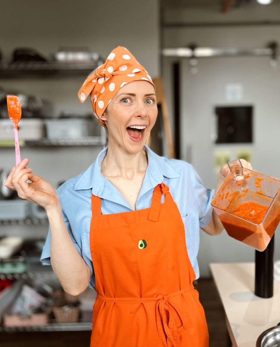 Fran Tonello de avental laranja e bandana de bolinhas, animada, com uma espátula e o copo do liquidificador nas mãos